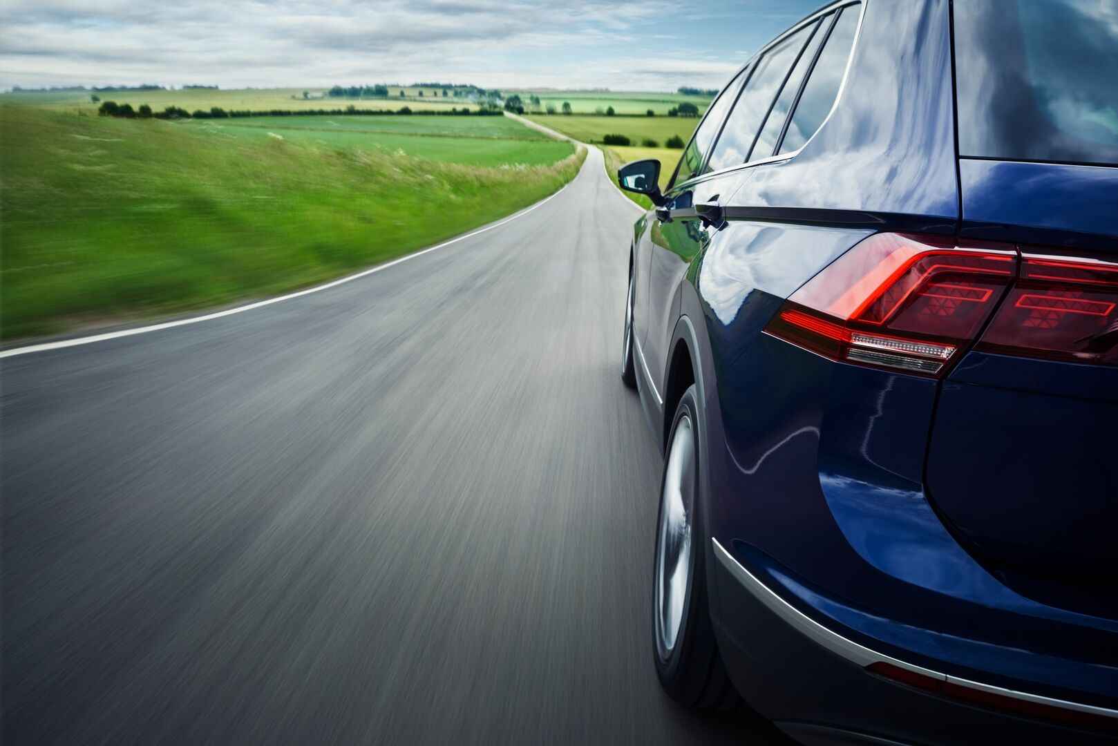 Close up shot of the back left side of a car driving down a road with countryside views in the distance